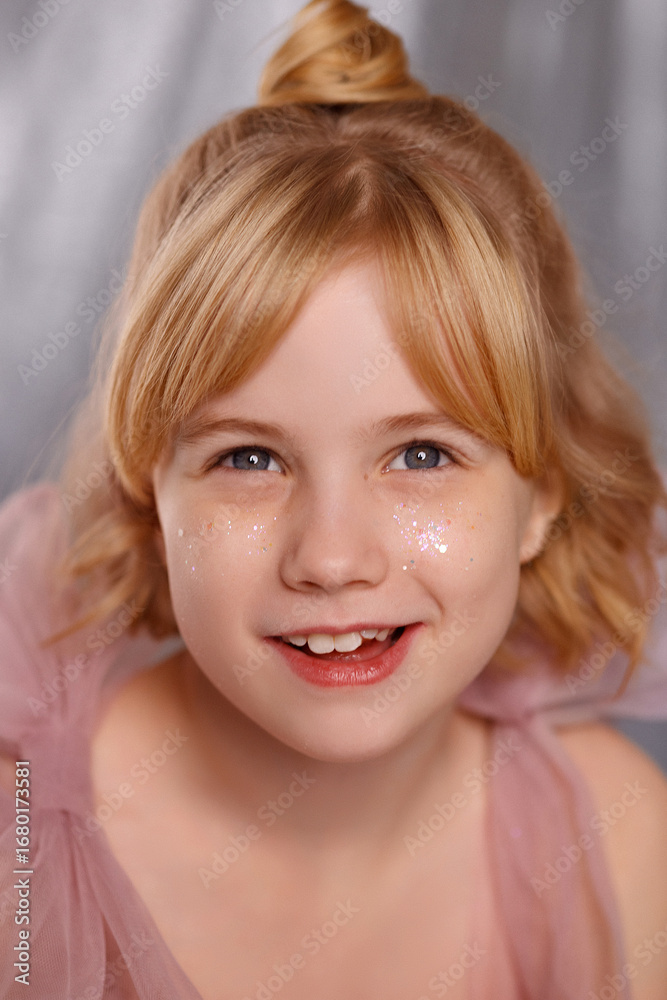 Naklejka premium Cheerful Caucasian girl, about 8 years old, blonde hair, pink dress, glitter on cheeks, front view, look at camera, gray background. Concept childhood, joy, celebration, innocence.
