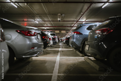 Bild auf Leinwand A dimly lit underground parking lot filled with cars parked in rows, featuring security cameras, concrete flooring, and overhead fluorescent lights