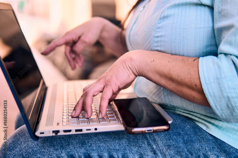 Naklejka premium View of the hands of an adult professional woman typing on a laptop, working online, connecting to the internet, and studying. She's using her cell phone, at home, outdoors. Planning, home office.