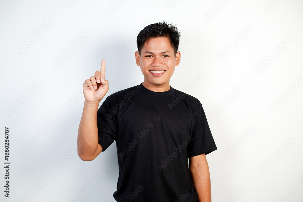 Fototapeta premium A Cheerful Young Asian Man Wearing a Black Shirt Pointing Upward With a Smile on a White Background.