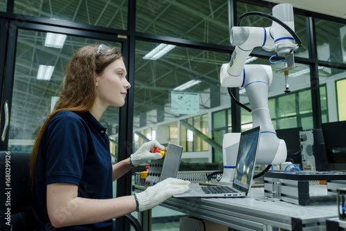 female robotics trainee sitting at smart factory workstation using control panel to manage robotic system during automation skill development training with laptop and robotic arm
