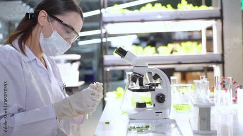 A female scientist wearing lab coat and mask conducts scientific experiments with microscope and test tubes in high tech lab.