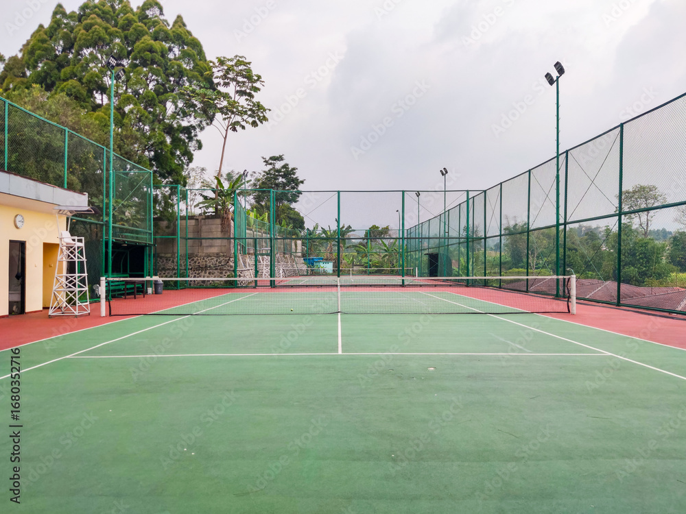 Fototapeta premium Empty tennis court with green and red surface, surrounded by tall wire fencing under an overcast sky, creating a quiet atmosphere for practice, competition, or leisure play in a peaceful outdoor setti