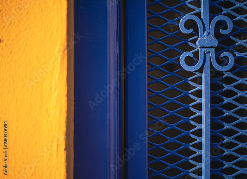 A striking contrast between a bright yellow wall and a deep blue metal grate is captured, showcasing architectural charm. The interplay of light and shadow highlights the textured surface of the wall 