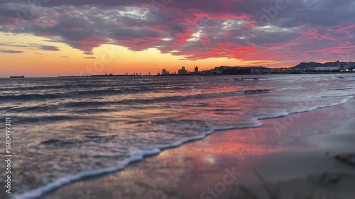A breathtaking sunset paints the sky with vibrant red, orange, and pink hues over the Durrës coastline. Waves gently wash onto the sandy beach, reflecting the colorful light of the sky.