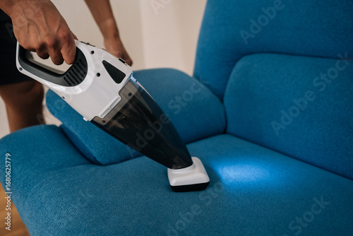 Close-up of cleaning service employee male using handheld vacuum cleaner to remove dust and dirt from blue sofa, emphasizing importance of cleanliness and hygiene in home maintenance.