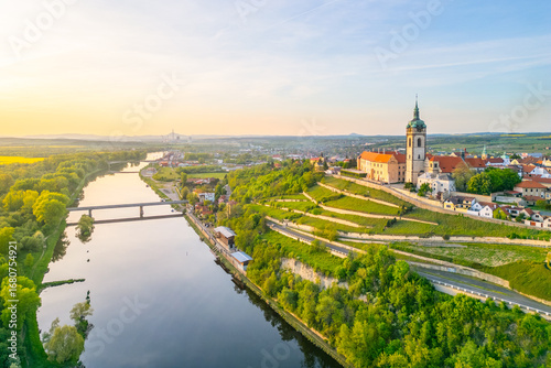 Aerial view captures Melnik City and its historic chateau perched above the serene Labe River. The landscape features lush greenery and tranquil waters during golden hour.
