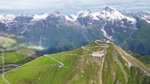 Aerial view of the winding Grossglockner High Alpine Road ascending a verdant mountain towards a structure, contrasting with snow-capped peaks, Austria.