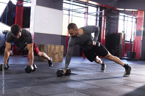 Diverse male training partners performing plank rows on mat in gym using hex dumbbells