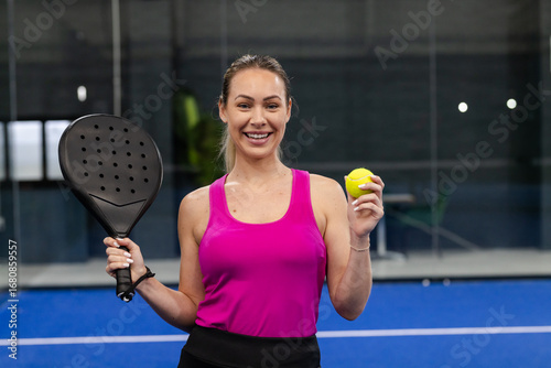 Woman standing on blue padel court in pink top, black skirt, holding racket, yellow tennis ball