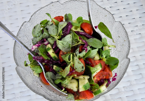 Fresh Cypriot salad in a glass bowl. A colorful, homemade salad with tomatoes, cucumbers, red cabbage, and fresh purslane leaves in a clear glass bowl, with two serving spoons.