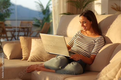 Happy woman at sunset at home using laptop on a sofa