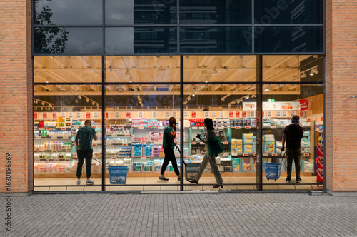 A look through a window in a supermarket at day time with people with people walking around shopping, looking at goods and talking on their mobile phones.