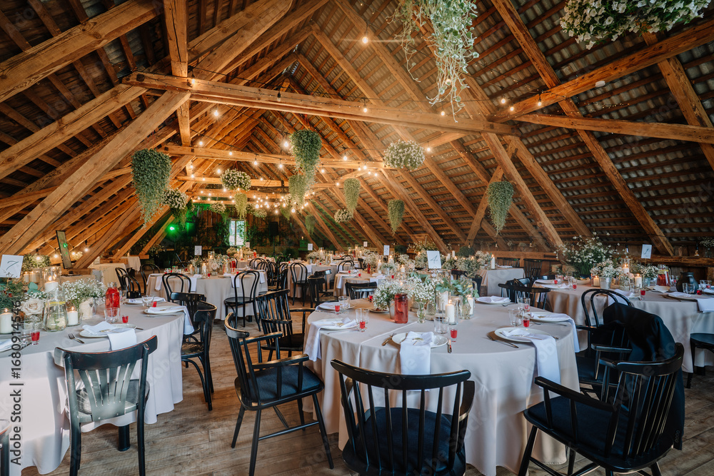 Fototapeta premium Rustic wedding reception setup in a wooden barn attic, decorated with fairy lights, floral arrangements, candles, and round tables covered in beige cloths.