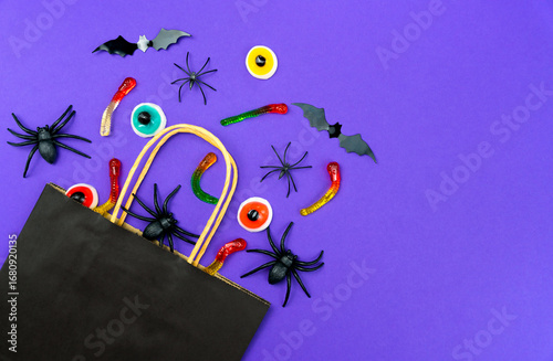 Black paper bag with holiday candies, gummy worms, toy spiders, bats and eyeballs on purple background. Halloween shopping and celebration concept. Top view. Copy space.