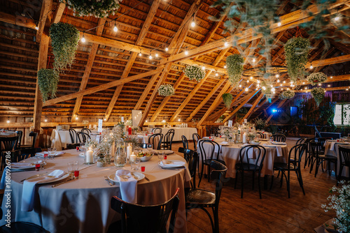 Fotografija A beautifully decorated rustic wedding reception hall with round tables, floral centerpieces, candles, wooden beams, and hanging greenery under warm lighting