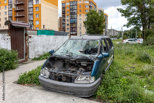 Wallpaper Mural Abandoned damaged car with shattered windshield and missing engine parts parked on overgrown grass near residential buildings.  Torontodigital.ca