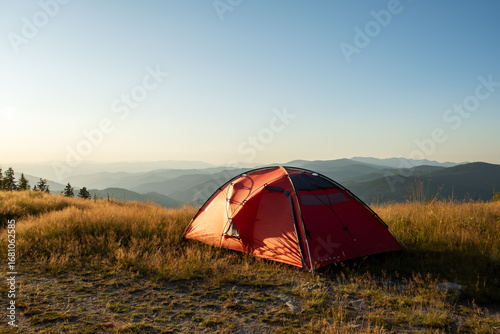 Morning mountain silence, red tent pitched on hill overlooking forested valleys, clean light and blue sky capture freedom and stillness of wild camping life, minimal gear, solo adventure story