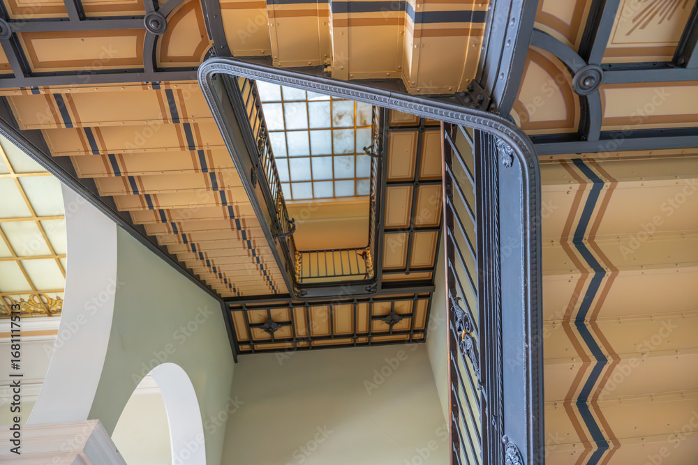 Obraz premium A decorated stairwell with patterned railings rises toward a frosted skylight. The ornate design creates a sense of elegance.