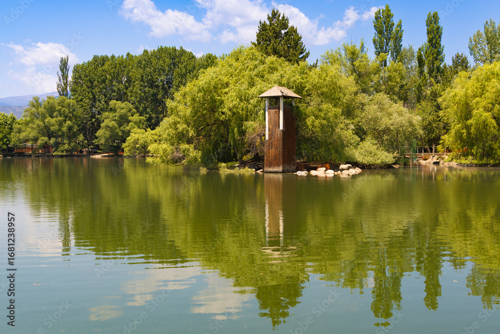 Obraz premium Wooden tower surrounded by lush greenery and calm waters at Schierbeck Park in Puigcerdà, with clear reflections and a bright blue summer sky.