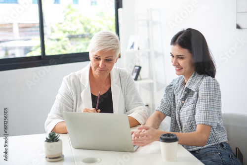 Schilderij op canvas Senior business women mentoring younger colleague with laptop in modern office, teamwork and professional collaboration concept for learning and corporate success