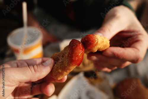 Two hands holding chicken nuggets with ketchup during picnic, fast food meal outdoors. Close up view