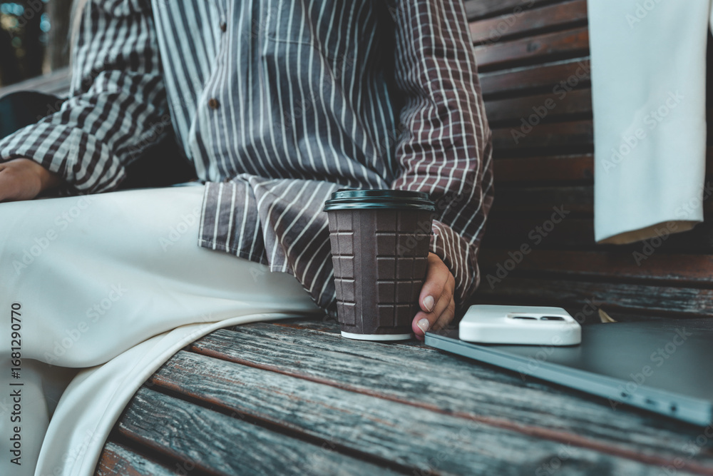 Fototapeta premium Close-up of coffee cup, smartphone, and laptop on wooden bench with woman’s hand holding the cup, cozy work and lifestyle moment outdoors