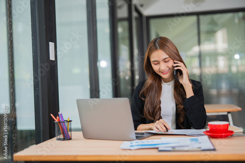  A beautiful businesswoman in a suit is looking at a computer screen while talking on her mobile phone. She is sitting in an office and using modern technology.