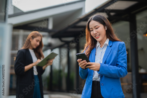 Asian businesswoman in suit using mobile phone while standing in front of modern business building. Free Download Asian businesswoman in suit using mobile phone while standing in front of modern business building. - GFXLoad