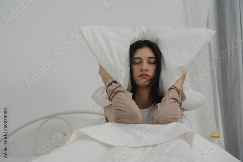 Fotografie Annoyed woman covering her ears with pillows while sitting in bed, suffering from noise disturbance, insomnia, or sleep problems