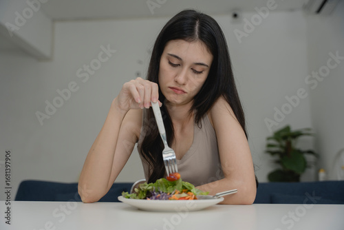 Young woman looking bored or unhappy while eating salad, pushing food with fork at table. Concept of appetite loss, diet frustration, healthy eating challenge, food aversion, or emotional eating issue