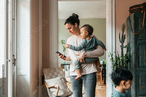 Woman wearing wireless in-ear headphones standing with baby boy while using smart phone at home