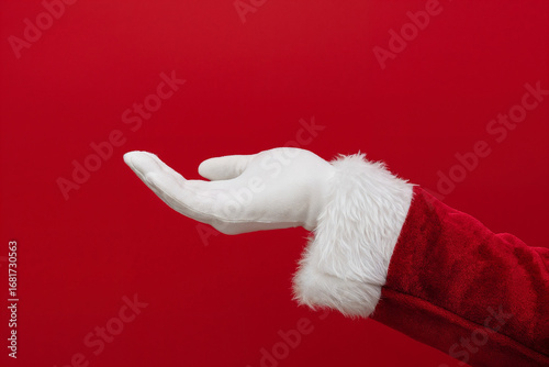 Santa Claus hand in white glove and red suit sleeve with fur trim reaching out, isolated on red background, symbolizing Christmas, gift giving, and festive holiday traditions.