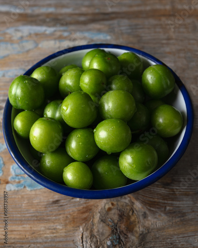 Close-up of fresh green plums in a white enamel bowl with blue rim, placed on a rustic wooden table.