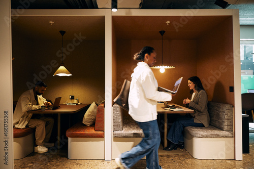 Female entrepreneur walking with laptop while colleagues sitting in hybrid coworking space at office