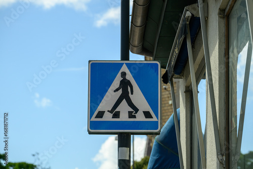 A prominent traffic sign shows a pedestrian icon on a blue background, alerting drivers to a nearby crossing. The structure is partially visible alongside the sign in bright daylight