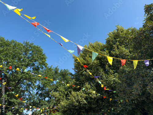 Colorful festive pennant flags hanging on strings against blue summer sky and green trees creating bright decorative background for holiday celebration and cheerful outdoor events