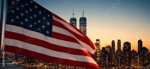 A patriotic view of the American flag waving proudly in the foreground with the iconic, illuminated New York City skyline and the Twin Towers visible in the background just before dusk.