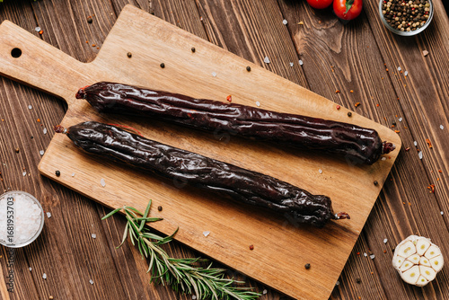 Fotografie Dry cured sausage sticks on wooden board with herbs, tomatoes, and spices