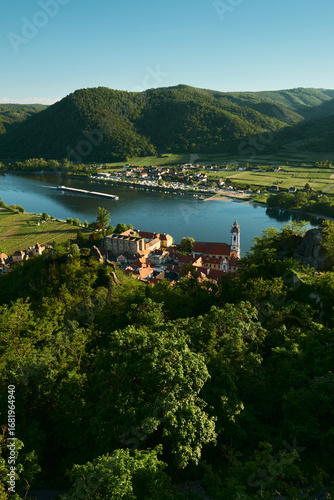 Vertical sunny view from above of a baroque church, historic town, forests, vineyards, hills, and the Danube River with a passing barge in Wachau Valley, Austria.