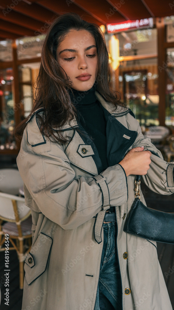 Fototapeta premium Portrait of stylish woman in beige trench coat and black turtleneck, holding a black handbag and gazing into the distance at a cozy outdoor café
