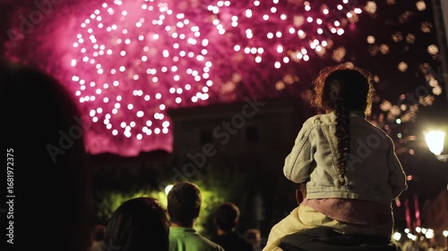 A little girl sits on her parent’s shoulders, watching dazzling fireworks illuminate the night sky. Magical festive atmosphere, childhood wonder, New Year, Christmas, holiday, festival, amazing