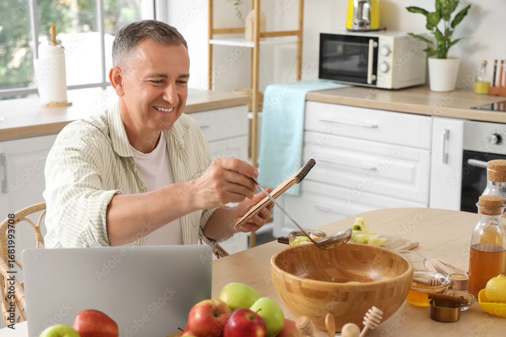 Fototapeta premium Mature man with recipe book and apple cider at table in kitchen