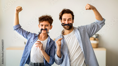 Fotografie Son and strong dad showing biceps, holding fake moustache on sticks and smiling to camera