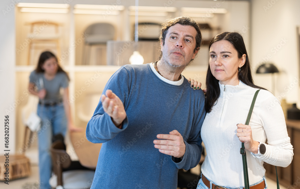 Fototapeta premium Husband and wife are discussing the purchase of new furniture for the house, they are considering furniture made of natural wood. European couple choose chairs and a sofa for an apartment after