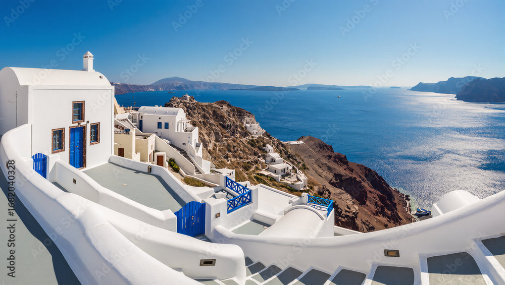 Obraz premium A panoramic view of the iconic white houses and blue domes of Santorini, Greece. The buildings are perched on a cliffside, overlooking the deep blue caldera and the Aegean Sea, with the sky clear.