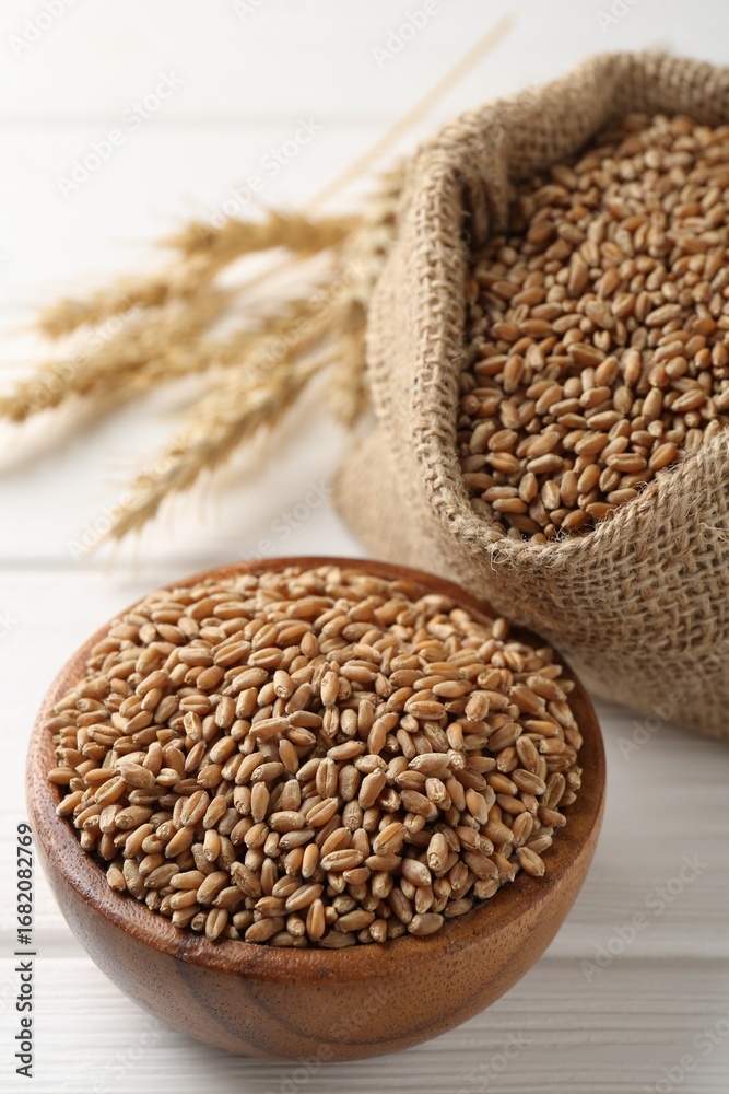 Fototapeta premium Wheat grains in bowl, burlap bag and spikelets on white wooden table, closeup