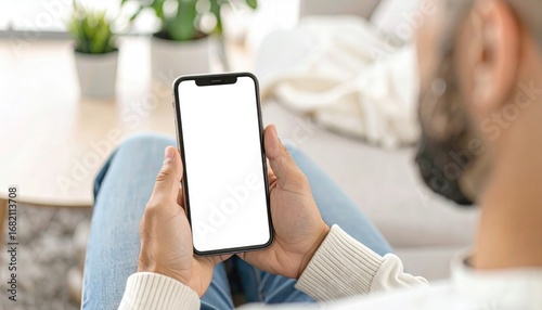 A man holds a smartphone with a blank white screen while seated on a couch. Plants and a table are visible in the blurry background