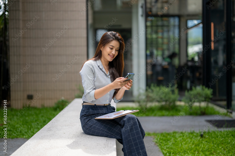 Fototapeta premium Smiling businesswoman using smartphone while sitting outdoors with documents