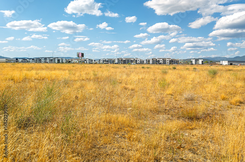 Rows of new construction townhomes and condominiums can be seen on the other side of a highway from a dry grass field on the American prairie.